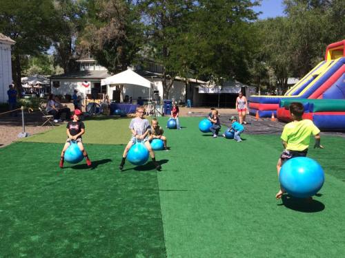 Children playing on blue bouncing balls on a grassy area, with inflatable structures and tents in the background.