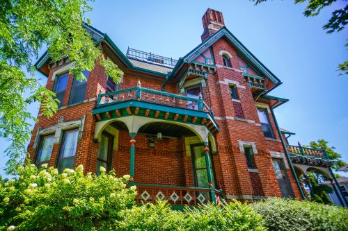 Victorian-style brick house with ornate details, green trim, and lush greenery in the foreground under a clear blue sky.