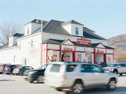 A white building with red accents houses a country store, surrounded by parked cars and a busy street.