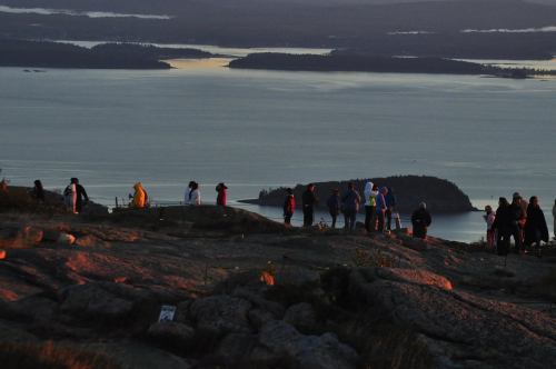 A group of people stands on a rocky hilltop, overlooking a serene lake and distant islands at sunset.