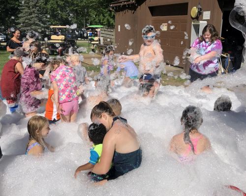 Children playing in a foam party, surrounded by bubbles and enjoying a sunny day at a park.