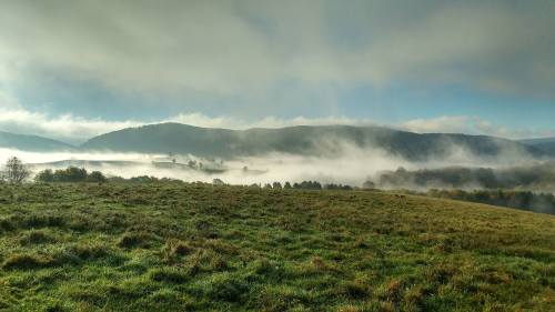 A misty landscape with rolling hills and a grassy foreground, under a blue sky with scattered clouds.