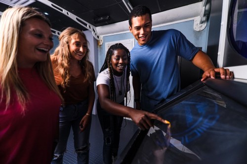 Four young people interact with a touchscreen display, smiling and engaged in a fun activity together.