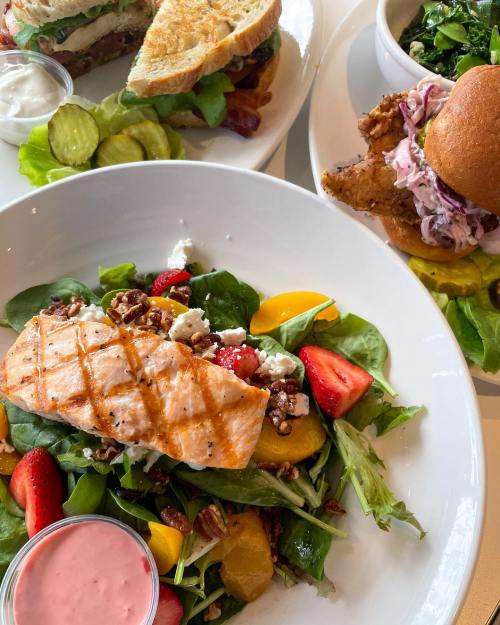 A vibrant salad with grilled salmon, strawberries, and pecans, served with a side of dressing and accompanied by sandwiches.