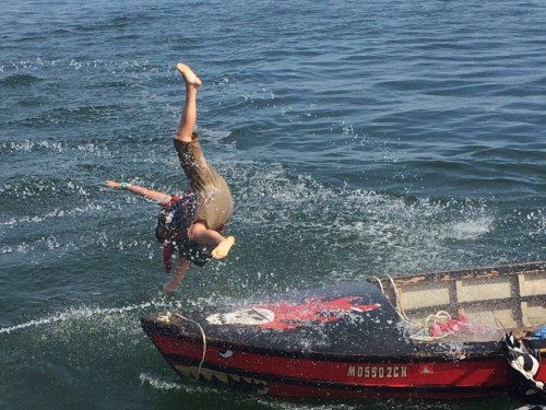 A person performs a handstand on the edge of a small boat, splashing water around in a sunny setting.