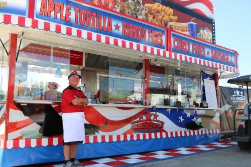 A vendor stands proudly in front of a colorful food stall featuring various snacks like apple tortillas and curly fries.