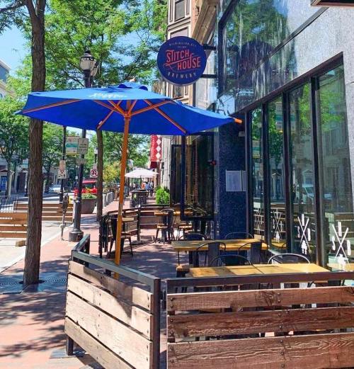 Outdoor seating area of a restaurant with blue umbrellas and wooden barriers, surrounded by trees and city buildings.