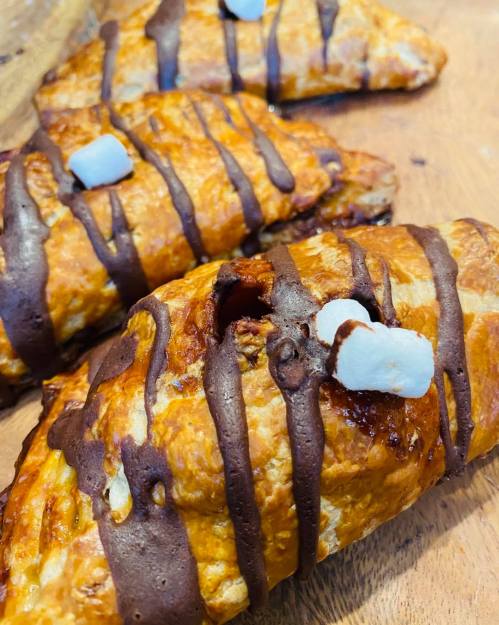 Three golden-brown pastries drizzled with chocolate and topped with small marshmallows on a wooden surface.