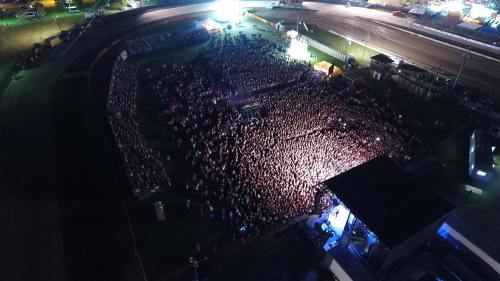 Aerial view of a large crowd at a nighttime outdoor event, with a stage and bright lights in the background.