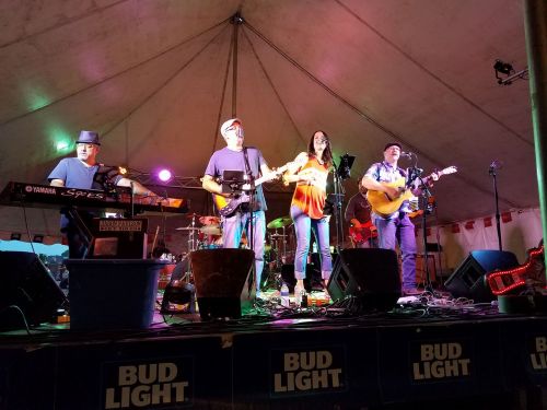 A live band performs on stage under a tent, with musicians playing various instruments and a singer in front.