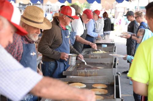 A group of people cooking pancakes at an outdoor event, wearing aprons and hats, with a crowd in the background.