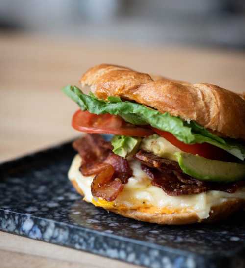 A close-up of a sandwich with bacon, lettuce, tomato, and cheese on a toasted bun, served on a dark plate.