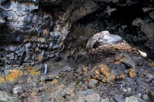 A rocky cave interior with textured walls and scattered stones on the ground, illuminated by natural light.