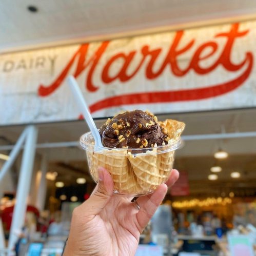 A hand holding a cup of chocolate ice cream topped with nuts, in front of a market sign.