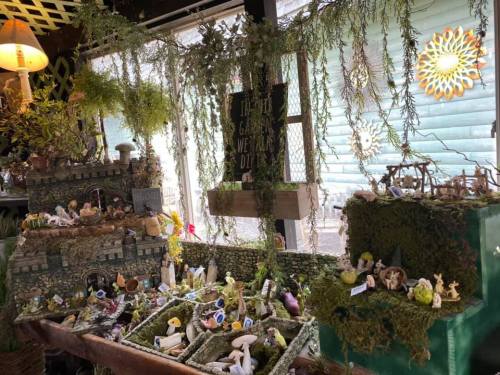 A whimsical display of miniature figures and decorations surrounded by moss and greenery in a cozy shop setting.