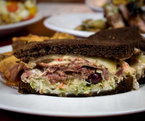 A close-up of a sandwich with layers of meat, cheese, and coleslaw, served on dark bread with sides in the background.