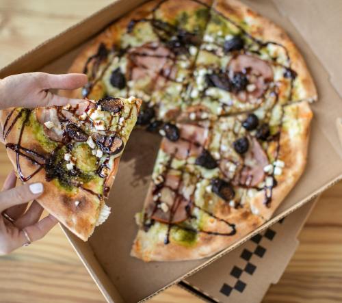 A hand holding a slice of pizza with toppings, next to a partially eaten pizza in a takeout box.