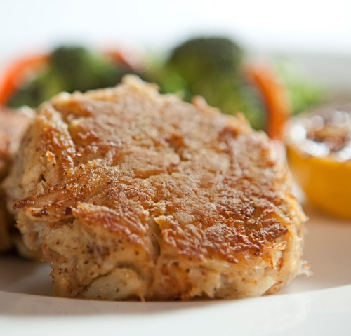Close-up of golden-brown crab cakes on a plate, accompanied by broccoli and colorful salad.