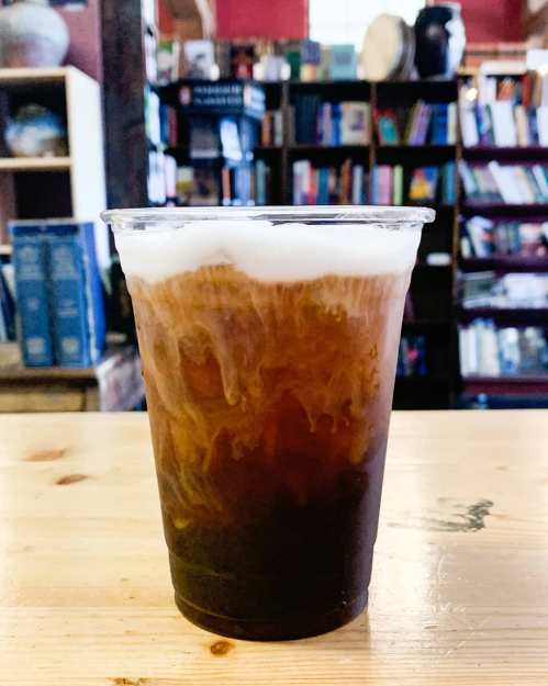 A layered drink in a clear cup, featuring dark coffee at the bottom and frothy cream on top, set on a wooden table.