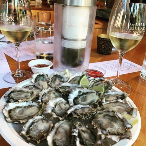 A plate of fresh oysters with lime wedges, accompanied by glasses of white wine and sauces on a wooden table.