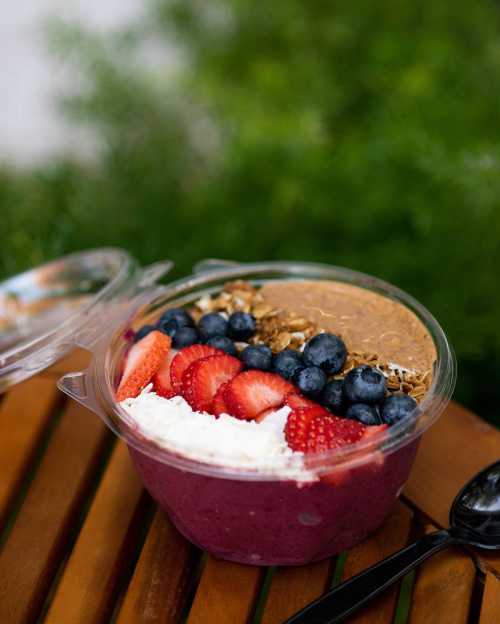 A colorful smoothie bowl topped with strawberries, blueberries, granola, and a dollop of nut butter, served in a clear container.