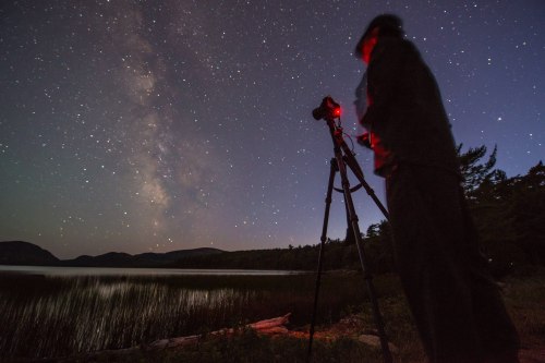 A silhouette of a person with a camera on a tripod, capturing the Milky Way over a calm lake at night.