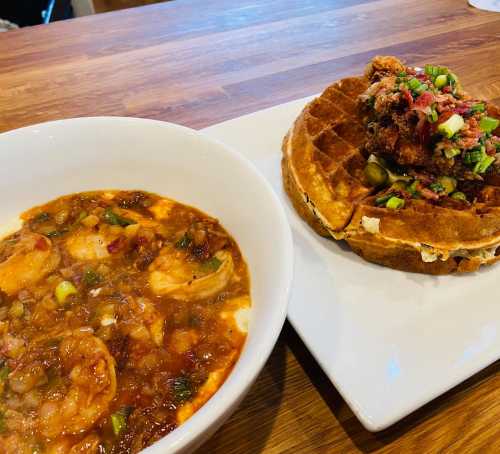 A bowl of shrimp stew next to a plate with a waffle topped with fried chicken and green onions.