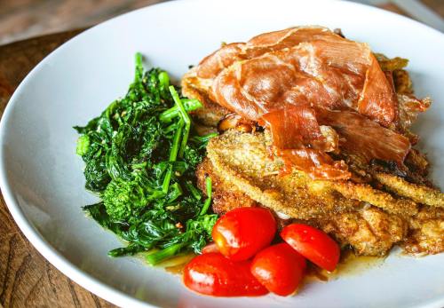A plate featuring crispy fried fish topped with prosciutto, sautéed greens, and cherry tomatoes.