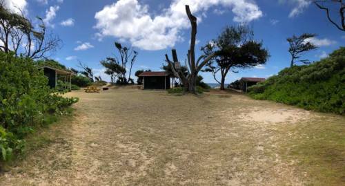 A grassy area with scattered trees and two small buildings under a partly cloudy sky.