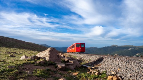 A red train travels along a rocky track with mountains and a blue sky in the background.
