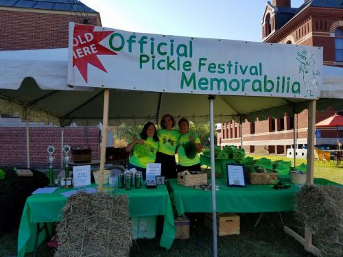 Three women in bright green shirts stand behind a table at a pickle festival, with a sign reading "Official Pickle Festival Memorabilia."
