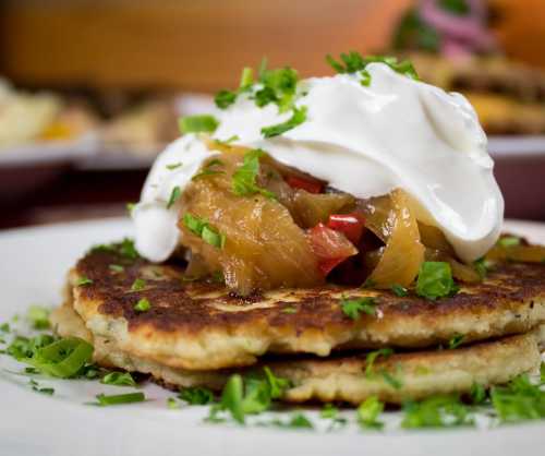 Close-up of pancakes topped with sautéed onions, red peppers, and a dollop of sour cream, garnished with fresh herbs.