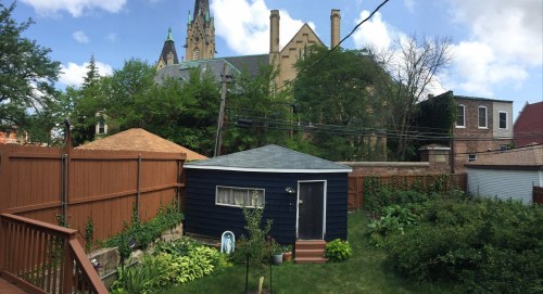 A backyard scene featuring a small blue shed, greenery, and a church steeple in the background under a cloudy sky.