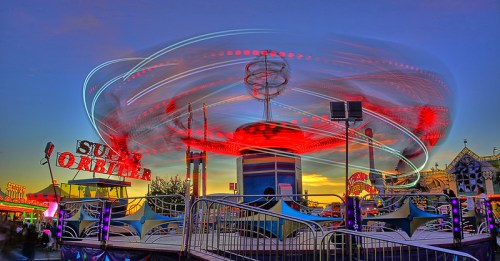 A vibrant carnival ride spins in the twilight, illuminated by colorful lights against a sunset backdrop.