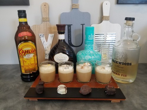 A display of various liquor bottles and four shot glasses filled with creamy drinks, alongside chocolate treats.