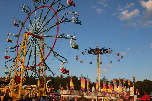 A vibrant carnival scene featuring a Ferris wheel and a swing ride, with colorful flags and a clear blue sky.