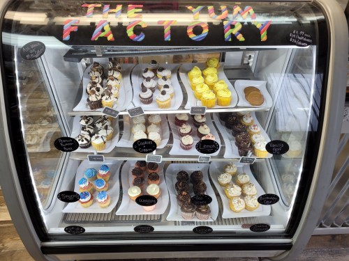 Display case filled with colorful cupcakes and desserts, labeled with flavors at The Yum Factory.