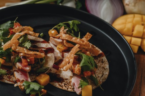 Two tacos topped with fresh vegetables, mango, crispy strips, and drizzled with sauce, served on a black plate.