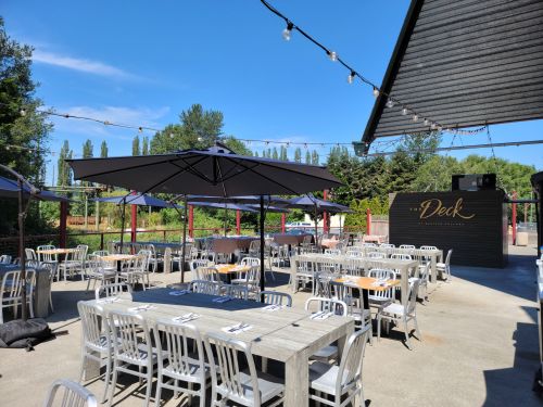 Outdoor dining area with metal tables and chairs, umbrellas, and a sign reading "The Deck" under a clear blue sky.
