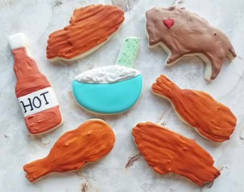 A variety of decorated cookies shaped like food items: chicken, a bottle of hot sauce, a bowl of rice, and a bison.