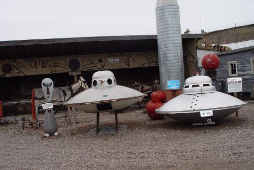 Three whimsical alien spacecraft and a cartoonish alien figure displayed outdoors, with a rustic backdrop.