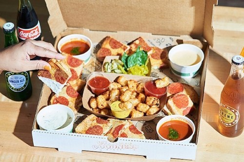 A pizza box filled with pepperoni pizza slices, dipping sauces, tater tots, and drinks on a wooden table.