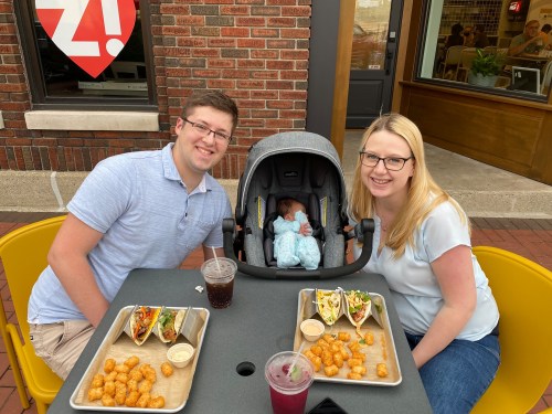 A couple sits at a table with food, smiling at the camera, while a baby sleeps in a car seat between them.