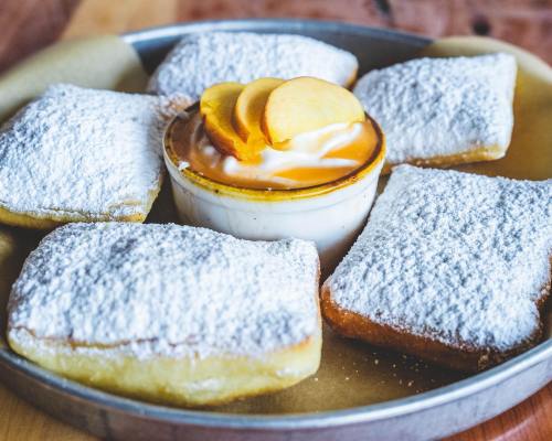A plate of five powdered sugar-covered beignets with a small bowl of caramel sauce and sliced fruit in the center.