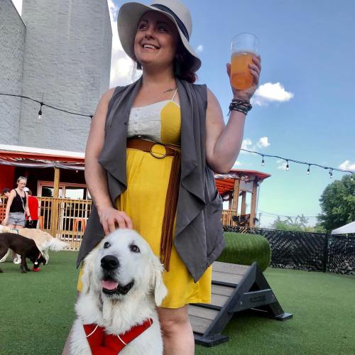 A woman in a yellow dress and hat holds a drink, standing next to a white dog on a grassy outdoor patio.