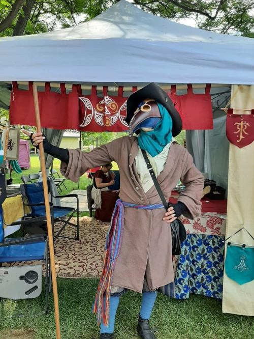A person in a bird mask and pirate-style outfit stands under a tent, holding a staff, with colorful decorations around.