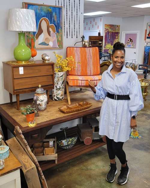 A woman in a striped shirt and black leggings stands beside a colorful chair on a display table in a vintage shop.