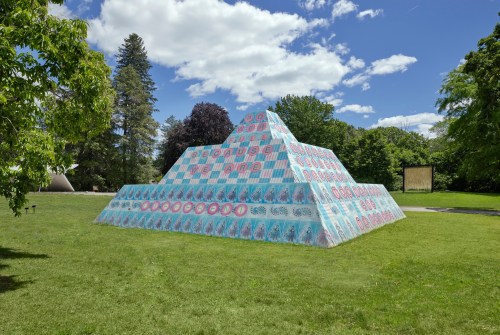 A colorful pyramid sculpture in a grassy park, surrounded by trees and under a blue sky with clouds.