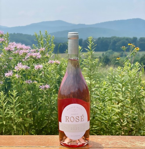 A bottle of 2020 rosé wine sits on a wooden table, with a scenic mountain view and wildflowers in the background.