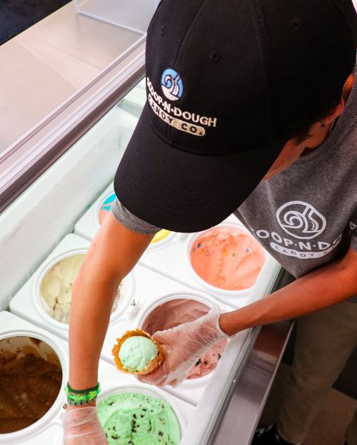 A person in a cap scoops mint ice cream into a cone from a display of various ice cream flavors.